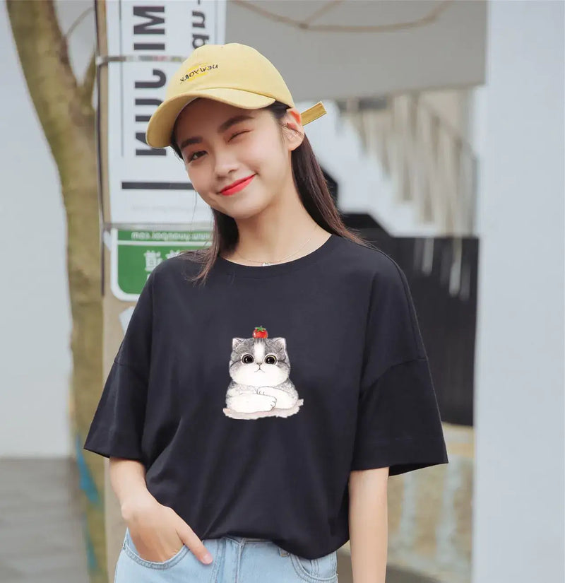 A woman wearing a black T-shirt with a cute cat graphic, smiling outdoors in a casual summer outfit.